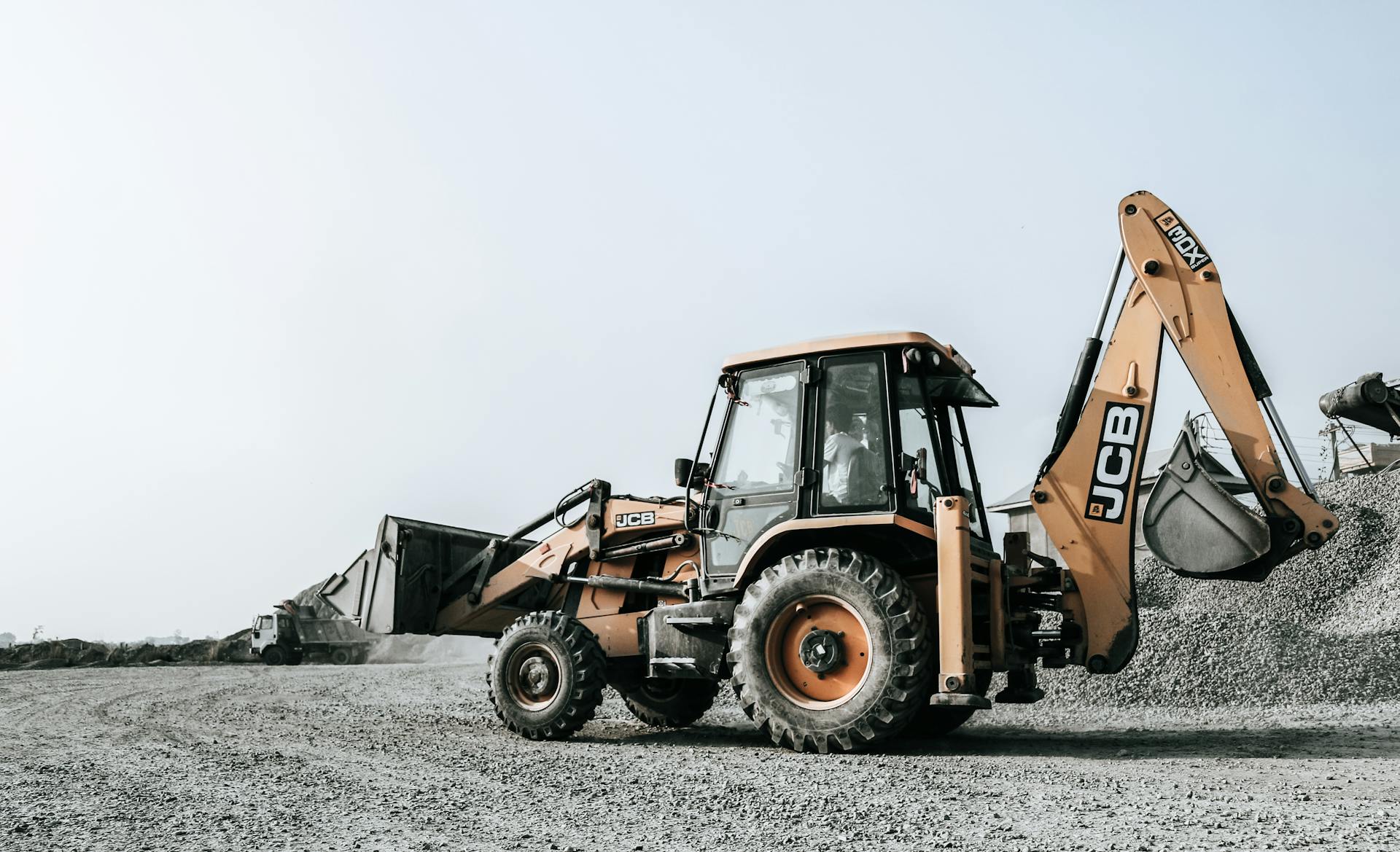 Widok z boku na żółtą koparko-ładowarkę marki JCB stojącą na placu budowy wysypanym żwirem. W tle widoczne są hałdy kruszywa pod jasnym, bezchmurnym niebem.